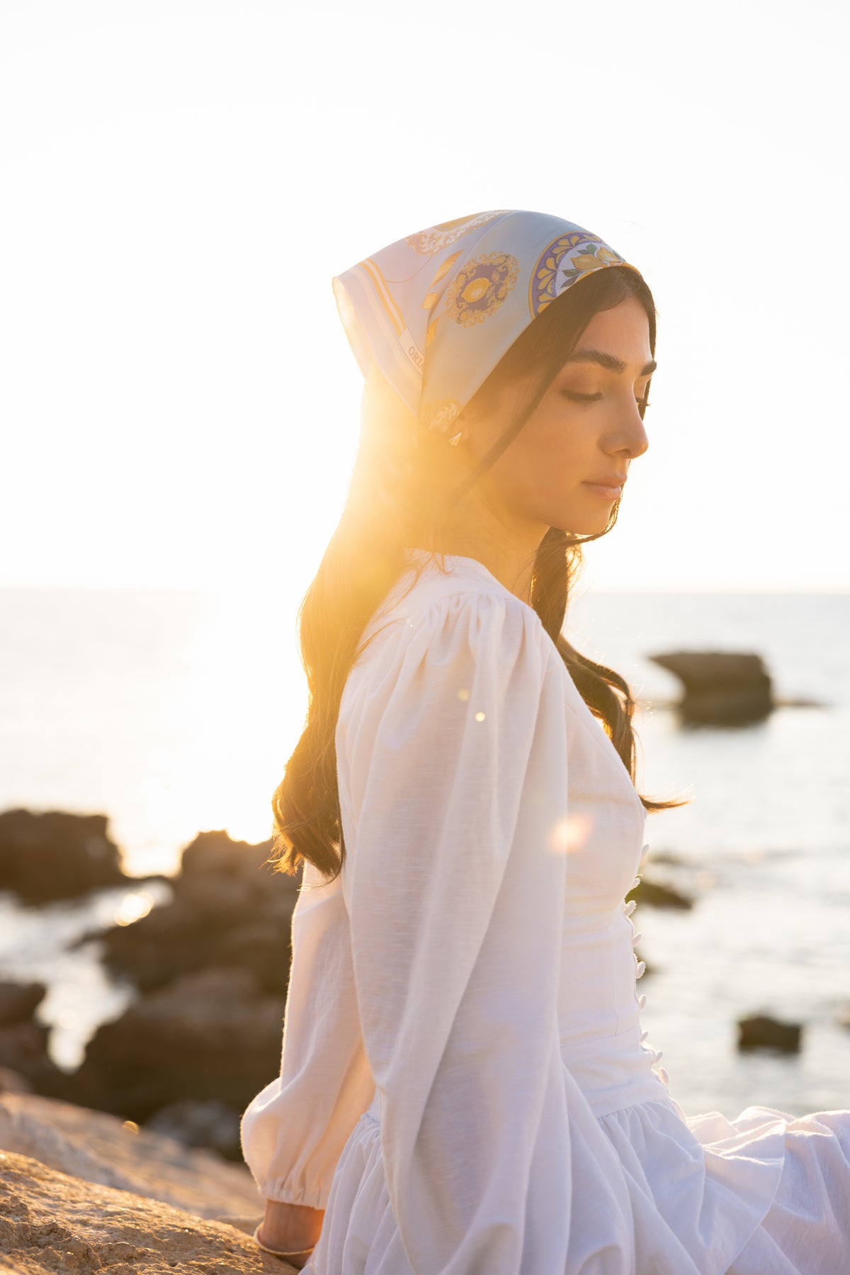 Foulard en soie Capri Eleganza bleu porté en bandana par une femme en robe blanche, assise sur les rochers au bord de la mer.