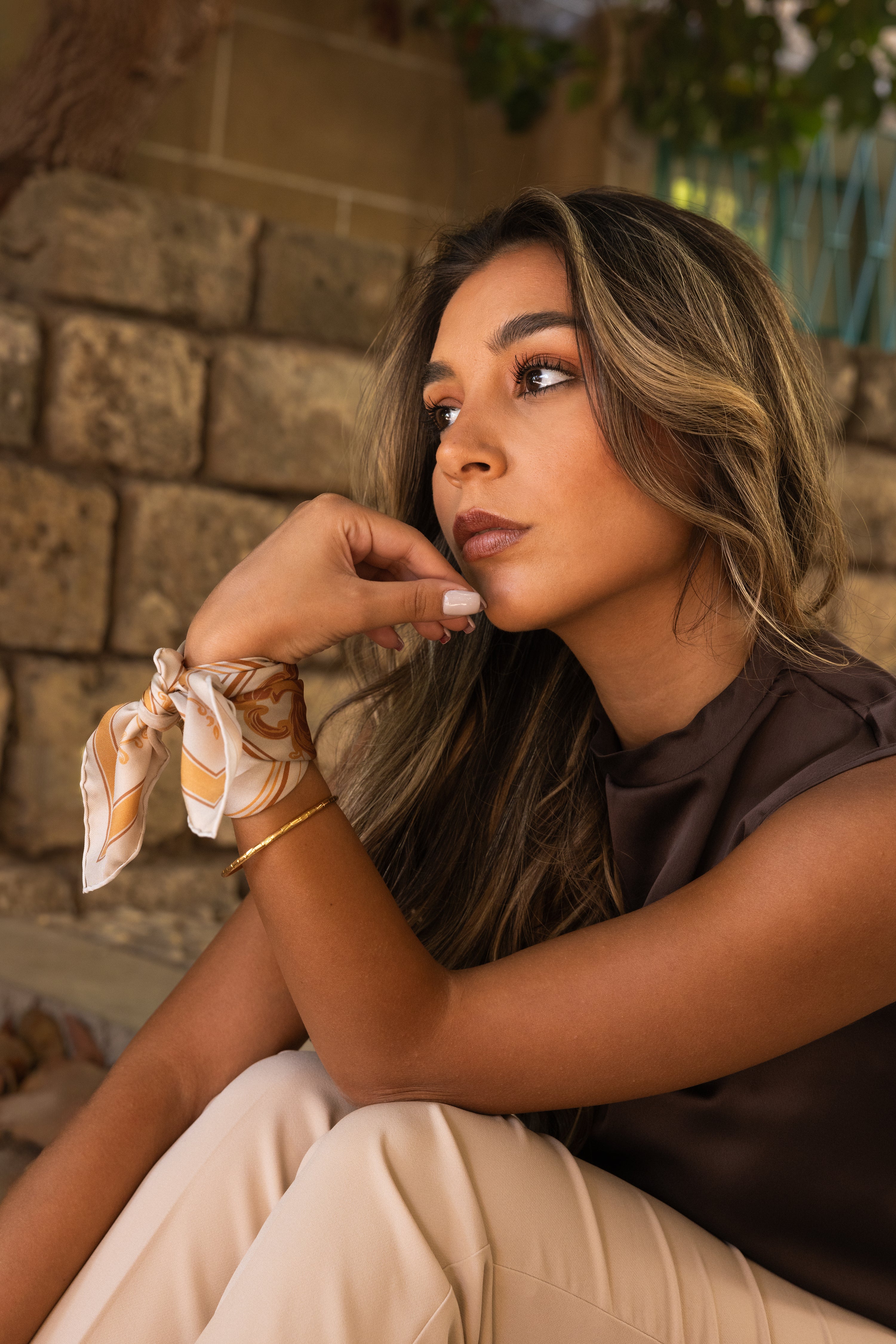 Vue sur le foulard Sole di Sicilia, porté au poignet, avec un bracelet en or en-dessous. Un mur en pierre apparaît à l'arrière plan.