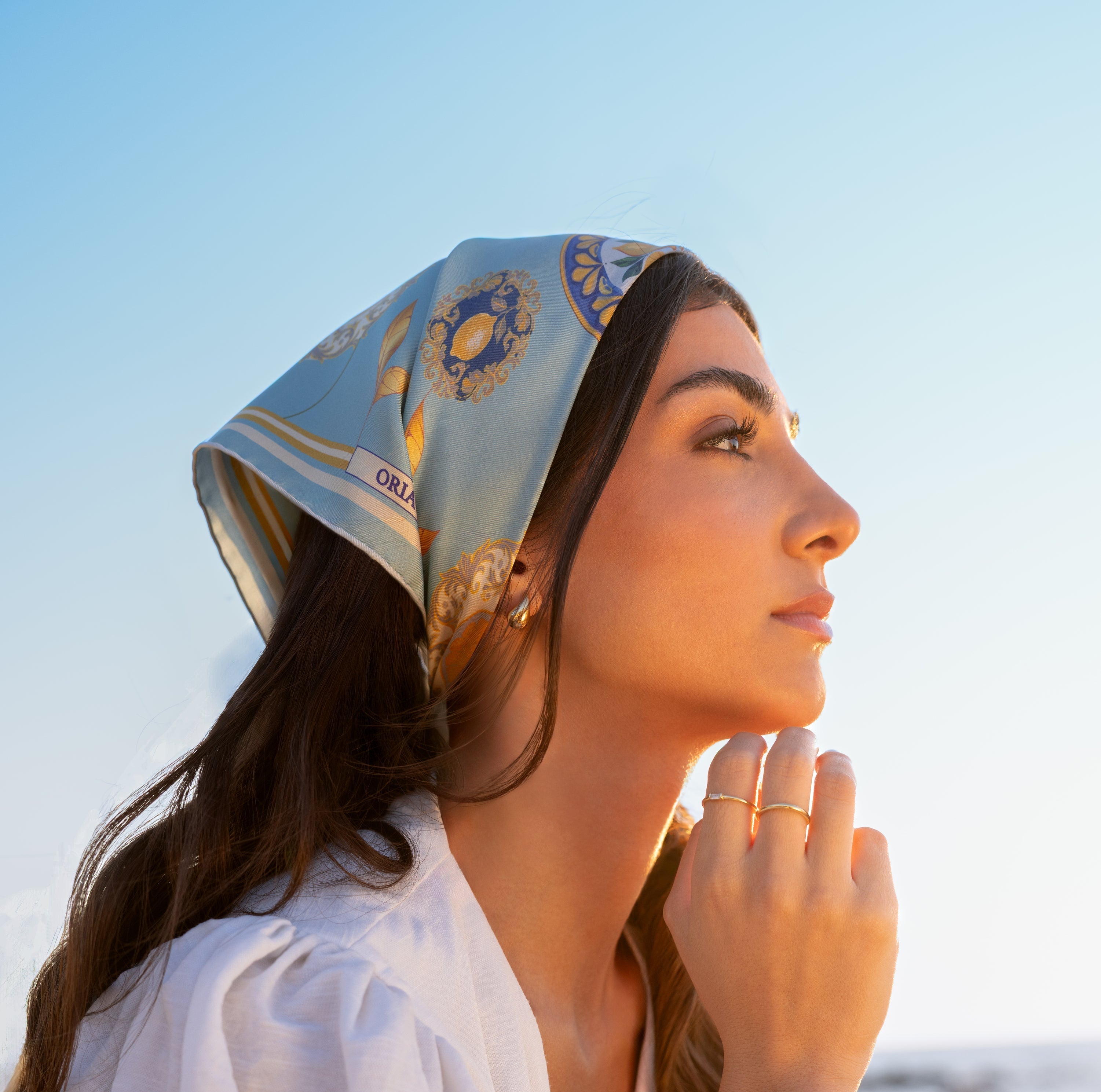 Photographie du foulard Capri Eleganza Bleu, porté sur la tête, avec des motifs de citrons, feuilles dorées et des références à la céramique de l'île, un ciel bleu en arrière plan.