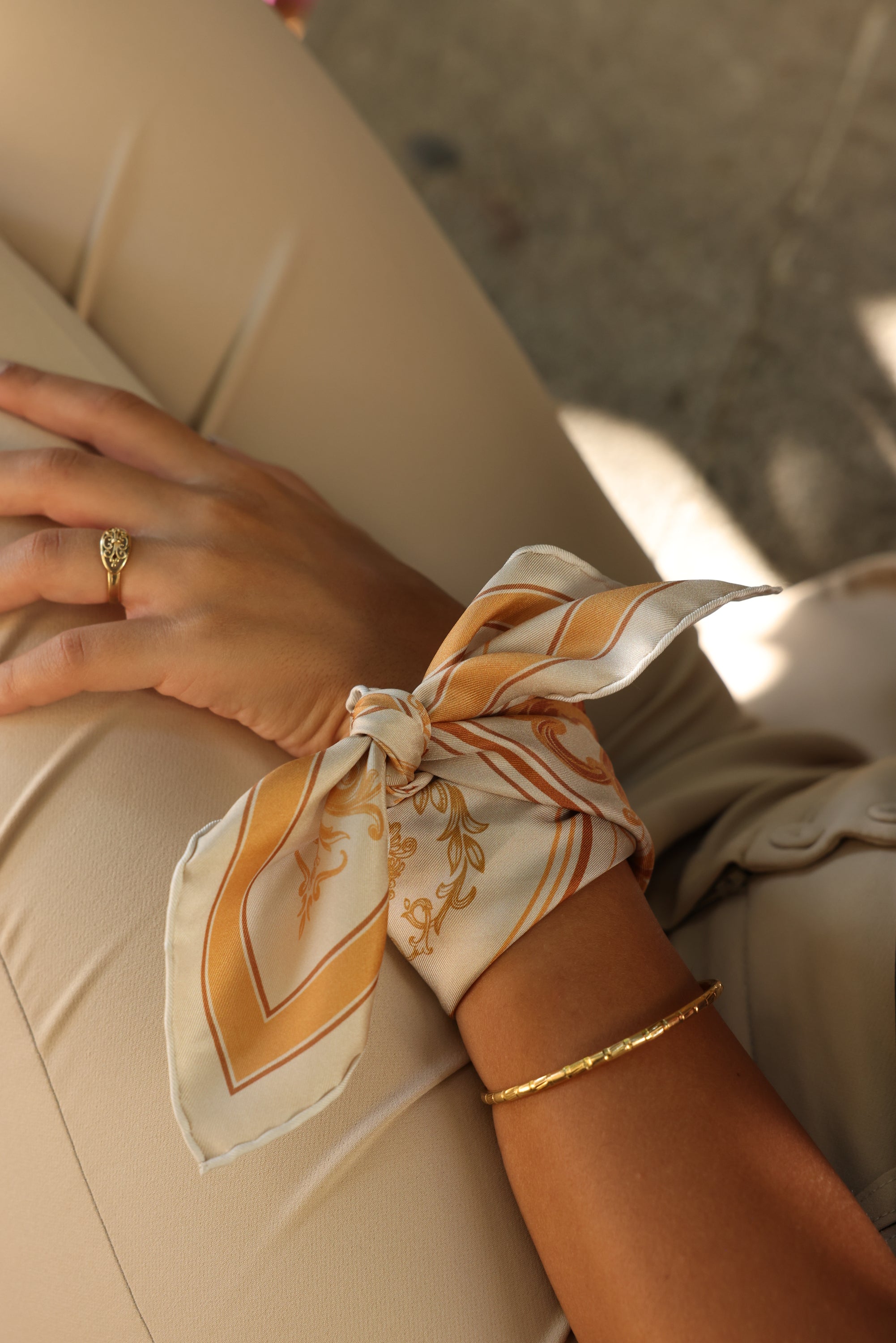 Vue du foulard Sole di Sicilia porté au poignet, avec un bracelet en or à côté et une bague en or au doigt, dans une ambiance lumineuse.